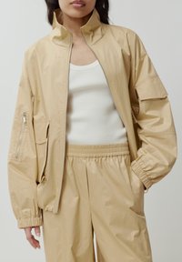 Beige Zip-Up-Jacke mit hohem Kragen, elastischen Bündchen und Seitentaschen. Getragen über einem weißen, gerippten Tanktop, kombiniert mit passenden beigen Hosen.