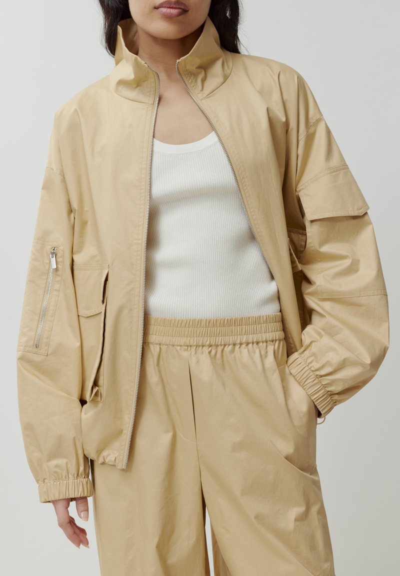 Beige Zip-Up-Jacke mit hohem Kragen, elastischen Bündchen und Seitentaschen. Getragen über einem weißen, gerippten Tanktop, kombiniert mit passenden beigen Hosen.