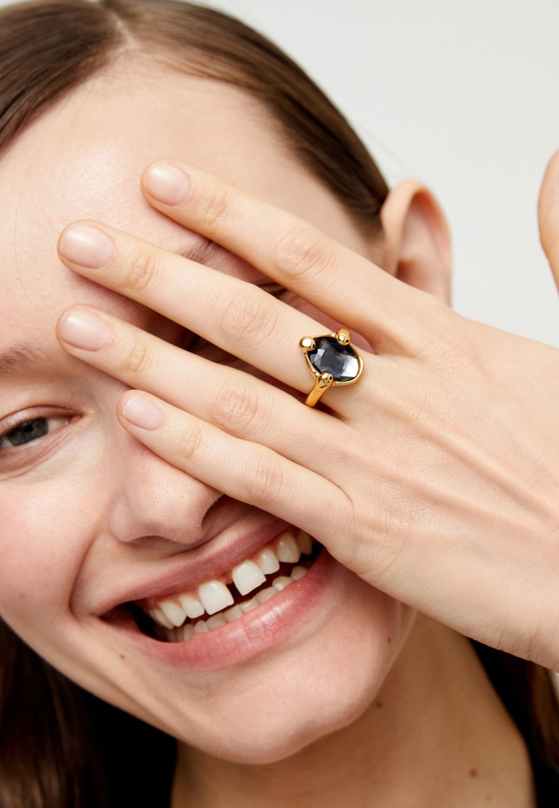 Anillo de oro con una gran piedra negra facetada y acentos redondeados, llevado en una mano que cubre parcialmente el rostro. Textura de piel suave.