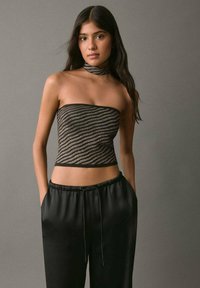 Top bez ramienok so čiernymi a zlatými pruhmi, ktorý má priliehavý strih, v kombinácii s voľnými čiernymi saténovými nohavicami s elastickým pásom a šnúrkou.