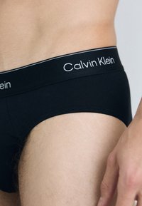 Schwarze Baumwollslips mit einem weißen „Calvin Klein“-Logo am Bund, die eine glatte Textur und eine taillierte Passform zeigen.