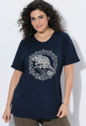 Vrouw die glimlacht en een marineblauw t-shirt met een gedetailleerd wit kameleon- en bladontwerp aan de voorkant draagt, staand tegen een witte achtergrond.