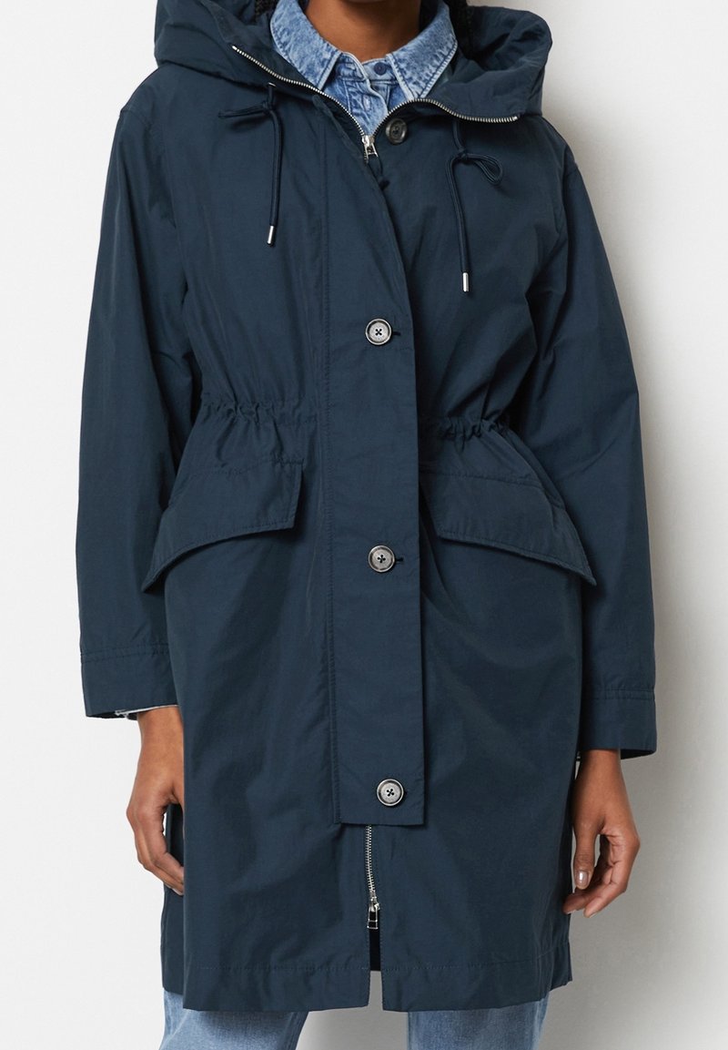 Parka à capuche bleu marine avec fermeture par boutons et fermeture éclair, taille ajustable, et poches à rabat à l'avant, portée sur une chemise en denim et un jean.