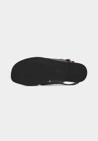 Μαύρος πάτος σανδάλου με λείο, επίπεδο κάτω μέρος και μερικώς ορατό λουράκι με χρυσή αγκράφα σε λευκό φόντο.