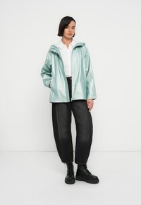 Πράσινο βροχερό μπουφάν μέντας με κουκούλα, κλείσιμο με κουμπιά snap, πλαϊνές τσέπες και λεία υφή, σε συνδυασμό με μαύρο φαρδύ παντελόνι και μπότες με κορδόνια.