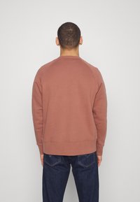 Sudadera de cuello redondo color burdeos con mangas raglán, textura suave y puños de canalé, combinada con jeans azul oscuro. Se muestra la vista trasera.