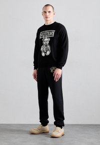 Sudadera negra con un gráfico de un oso blanco y el logo “Moschino”, combinada con pantalones negros que cuentan con un pequeño detalle de oso dorado, y zapatos beige.