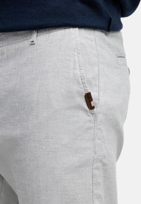 Pantalones de lino gris claro con un acabado texturizado, que cuentan con un parche de cuero marrón con el logo, bolsillos laterales y un corte entallado.
