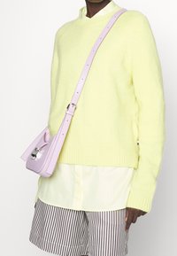 Hellgelber, flauschiger Strickpullover mit geripptem Saum. Kombiniert mit einem cremefarbenen Hemd und braun gestreiften Shorts. Kleine lavendelfarbene Umhängetasche mit silbernen Beschlägen.