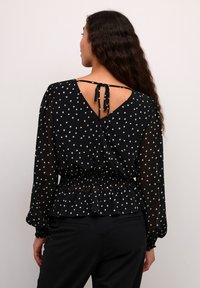 Blusa nera con pois bianchi, in tessuto trasparente, schiena a V con chiusura a fiocco, maniche lunghe a sbuffo e dettaglio in vita arricciato.