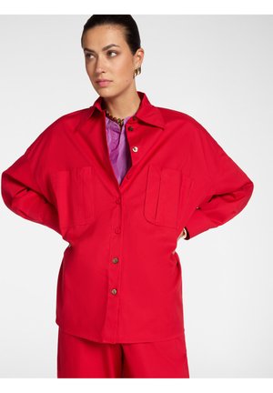 Frau mit dunklen Haaren trägt ein übergroßes rotes Hemd mit Knöpfen und passende Hose, darunter ist ein lila Top sichtbar.