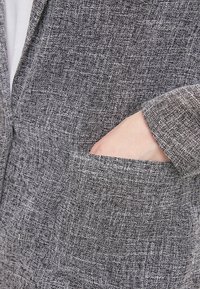 Tissu gris texturé avec un motif de tissage subtil ; présente une petite poche latérale et un détail de manche avec revers. Gros plan sur le vêtement.