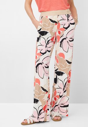 Femme portant un pantalon à jambes larges avec un grand imprimé floral beige, rose, noir et corail, assorti à des sandales blanches et un top corail.