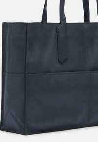 Granatowa skórzana torba typu tote z dwoma górnymi uchwytami, o zwartej konstrukcji, gładkiej fakturze i subtelnym logo z boku. Posiada jedną zewnętrzną kieszeń.