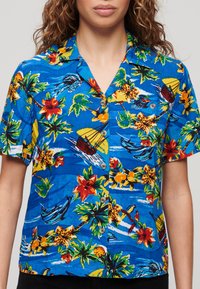 Camisa de manga corta azul con un patrón floral tropical en tonos amarillos, rojos y verdes, y botones marrones. Tejido ligero.