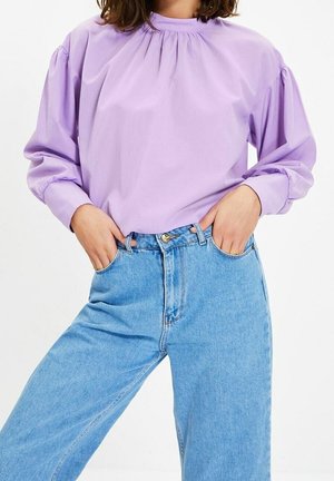 Nainen, jolla on päällään pitkähihaisen vaaleanvioletin pusero, jossa on kerätty pääntie, ja vaaleansiniset korkeavyötäröiset farkut, kädet taskuissa.