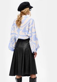 Vrouw met golvend blond haar in een paardenstaart, gekleed in een blauw-witte gebloemde blouse, zwarte leren rok, zwarte laarzen, een zwarte pet en een zonnebril.
