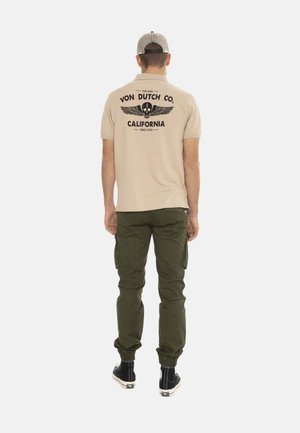 Homme portant un polo beige Von Dutch Co. avec un logo tête de mort et ailes, un pantalon cargo vert olive, des baskets montantes noires et une casquette grise, vue de dos.