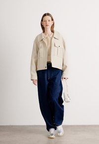 Beige cropped takki, jossa on kaksi etutaskua, päällä vaalea toppi ja tummat väljät farkut. Mallilla on päällään hopeiset lenkkarit ja hän pitää olkalaukkua.