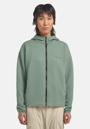 Person wearing a light green textured full-zip hooded jacket and beige pants, standing against a plain light background.