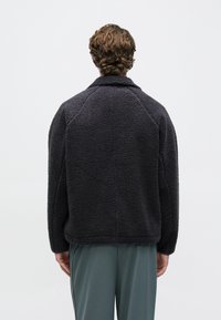 Χνουδωτό, σκουρόχρωμο γκρι φλις μπουφάν με γιακά και ραγκλάν μανίκια. Διαθέτει εμφανείς ραφές και άνετη εφαρμογή, φορεμένο πάνω από πράσινο παντελόνι.