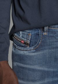 Vaqueros de mezclilla azul con un acabado desgastado, con costuras naranjas, una etiqueta negra y un diseño ajustado. Acompañados de una camiseta de manga larga gris.