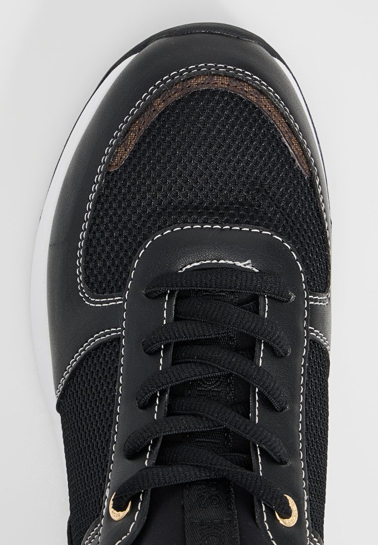 Zapatilla negra con tejido de malla, cordones negros, costuras blancas y un acento marrón en la punta, vista desde arriba sobre un fondo blanco.