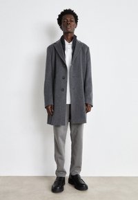 Cappotto grigio in misto lana con colletto a revers, sette bottoni e tasche laterali, abbinato a una polo bianca e pantaloni grigi. Scarpe nere completano l'outfit.