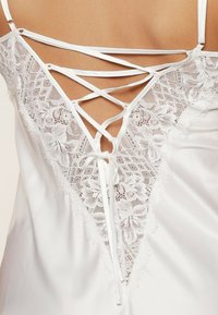 Abito in satin bianco con pannello in pizzo a motivi floreali, spalline incrociate sul retro e un dettaglio a fiocco sul fondo schiena.