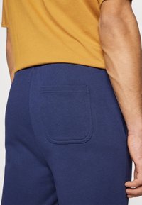 Námořnické jogger kalhoty s jednou zadní kapsou, vyrobené z měkké látky. Pas je elastický pro pohodlí, zobrazené s hořčicovým topem.