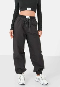 Pantalones negros de corte suelto con cintura elástica y cordón, tela texturizada, con puños en los tobillos, combinados con zapatillas deportivas blancas.