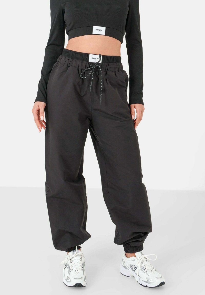 Pantalones negros de corte suelto con cintura elástica y cordón, tela texturizada, con puños en los tobillos, combinados con zapatillas deportivas blancas.