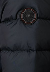 Granatowa puchowa kurtka z okrągłym czarnym logo z czerwonymi akcentami. Gładki, błyszczący materiał o pikowanej teksturze z widocznym przeszyciem.