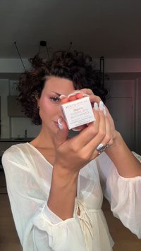 Revolution Eye Bright Corrector i en firkantet hvit beholder med rosa tekst, holdt i en hånd. Lokket er litt av, og viser produktet inni.