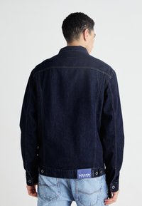 Mörk denimjacka med krage, synliga sömmar och knäppte ärmslut. Har en blå logotyp-patch vid midjan bak.