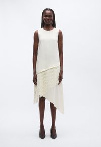 Rochie crem asimetrică cu un strat superior neted și partea inferioară cu franjuri. Are un decolteu rotund și este combinată cu pantofi cu toc negri.