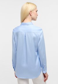 Blusa in raso azzurro chiaro con colletto, maniche lunghe e una piega centrale sul retro. Presenta polsini con bottoni e una superficie liscia.