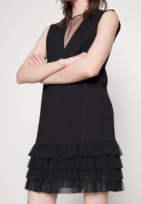 Sort ærmeløs kjole med gennemsigtigt mesh-halsudskæring og lagdelt tylvolantkant. Stoffet virker glat med en struktureret pasform.