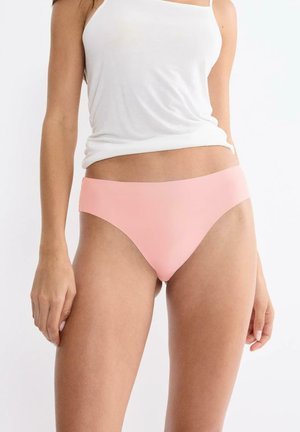 Woman wearing light pink seamless bikini underwear and a white sleeveless top against a plain white background.