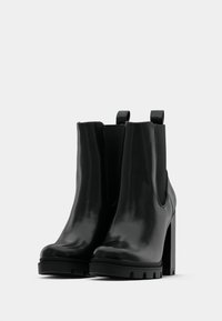 Un par de botines de cuero negro con tacones altos y bloques, y lengüetas para facilitar el uso, sobre un fondo claro y liso.