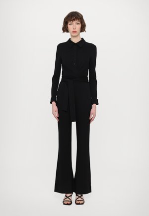 Zwarte, lange-jumpsuit met een kraag, knoopdetails, wikkelontwerp en een ceintuur in de taille. Kenmerkt zich door uitlopende broekspijpen en sandaaltjes met banden.
