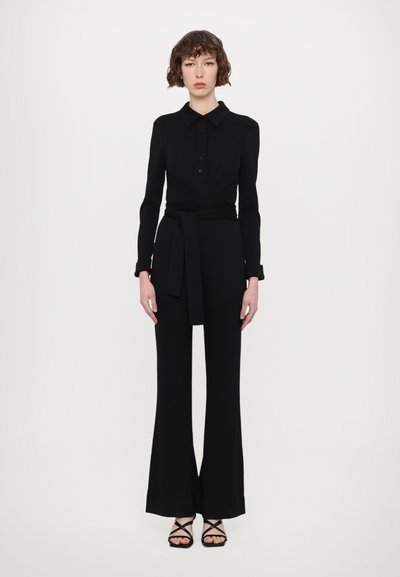 Black, long-sleeve jumpsuit with a collared neckline, button detailing, wrap design, and a belted waist. Features flared pant legs and strappy sandals.