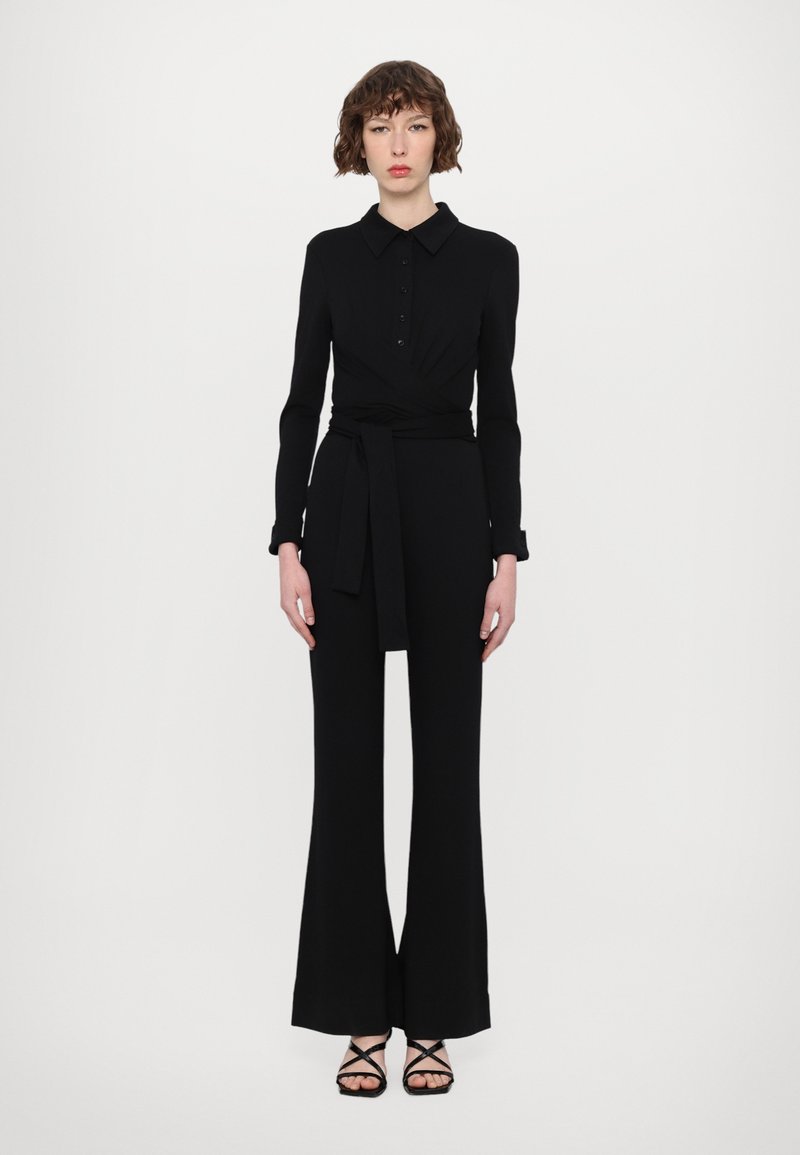 Combinaison noire à manches longues avec un col, des détails de boutons, un design enroulé et une taille ceinturée. Présente des jambes de pantalon évasées et des sandales à lanières.