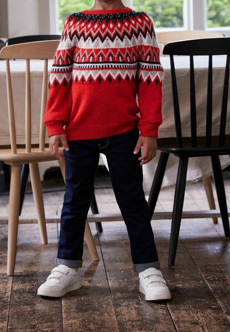 Enfant portant un pull à motifs rouges, blancs et noirs, un jean foncé et des baskets blanches, debout à l'intérieur entre des chaises en bois sur un sol rustique.