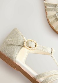 Glittery gold sandals with a textured finish, featuring multiple straps and a round buckle adorned with faux pearls. Flat, light brown sole.