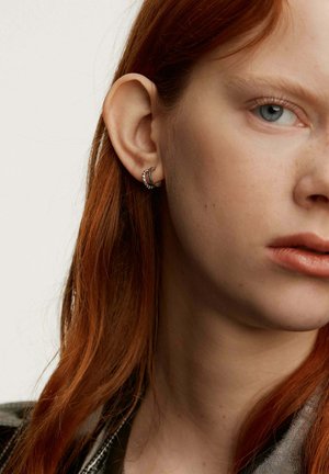 Pendientes en forma de aro plateados con una superficie texturizada, que presentan hilos entrelazados, llevados en la oreja de un modelo. El tono de piel es claro con sutiles pecas.