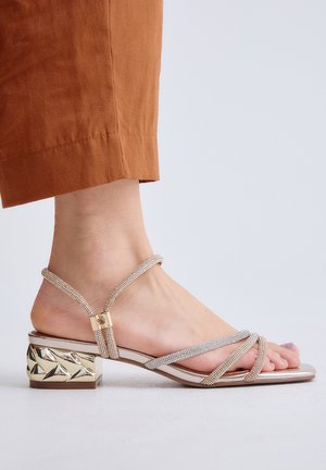 Pie con una sandalia dorada con tiras delgadas y brillantes y un tacón bloque con un diseño metálico y texturizado, combinado con pantalones color óxido.