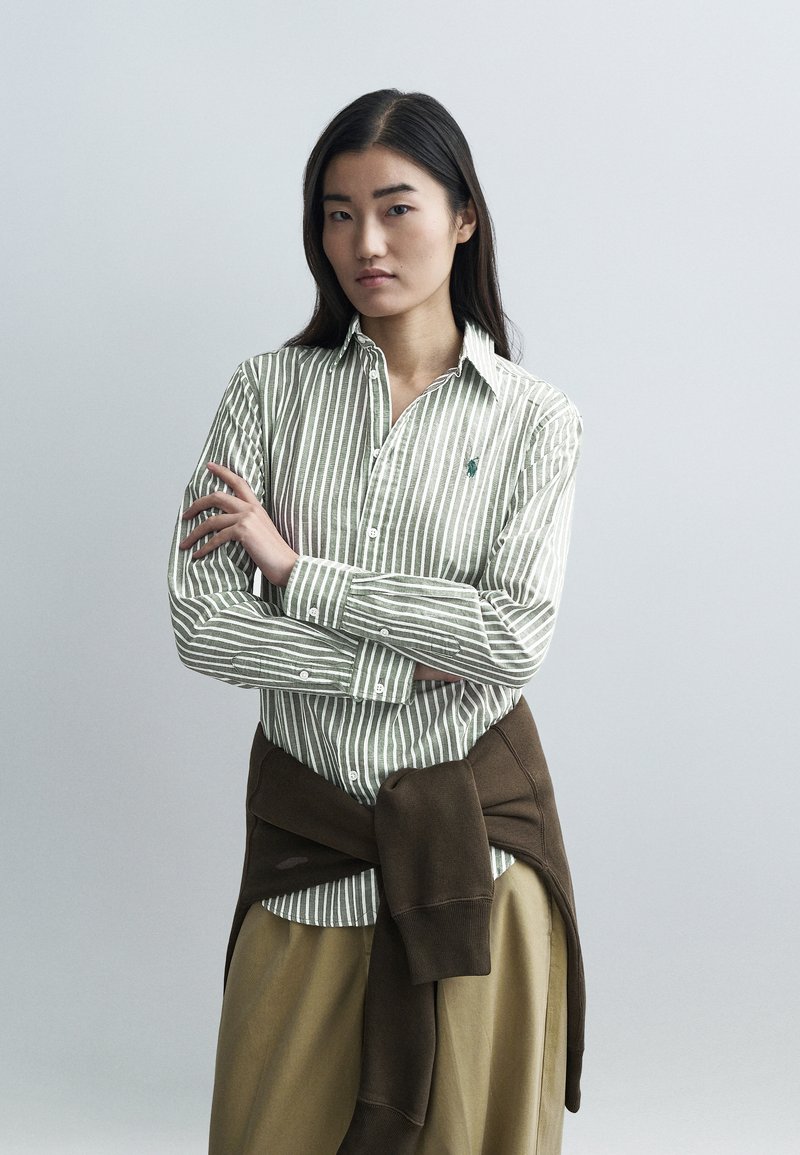 Zelená a biela pruhovaná bavlnená košeľa s gombíkmi a dlhými rukávmi, doplnená hnedým svetríkom zviazaným v páse.
