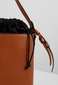 Sac seau en cuir marron avec fermeture en tissu à cordon noir et bandoulière réglable avec accents argentés.