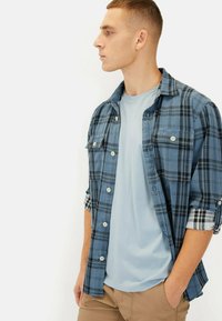 Blåternet button-up skjorte over en lys blå t-shirt, med en afslappet pasform og oplyste ærmer, parret med khaki bukser.